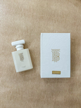 A perfume bottle and its matching box from the brand Thomas Blonde displayed. The bottle is a frosted off-white glass with a white cap, and the box is white with a gold monogram logo and a small gold brand plaque.