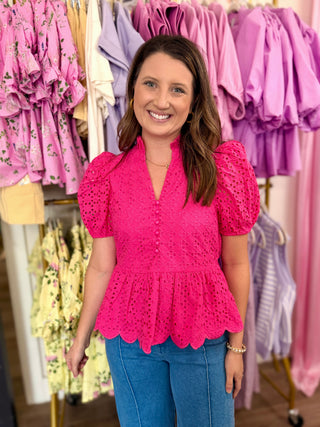Woman wearing a hot pink eyelet detail top in a clothing store with racks of colorful dresses.