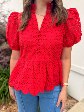 Person wearing a red eyelet detail top with puffed sleeves and blue jeans.
