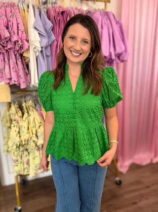 Woman wearing a green eyelet detail top in a clothing store with racks of colorful dresses in the background.