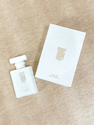 A white rectangular perfume bottle with a matching white box, both featuring a gold monogram logo