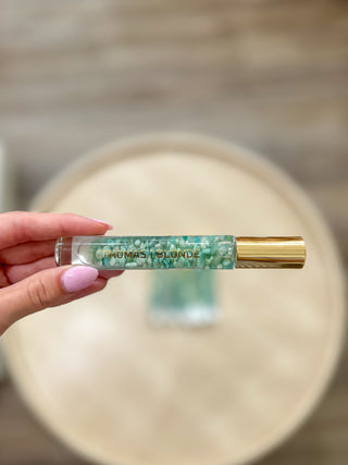 A clear glass rollerball bottle containing green and white crystals inside the liquid. The bottle has a shiny gold cap and the brand name 'THOMAS BLONDE' is printed in gold on the front.