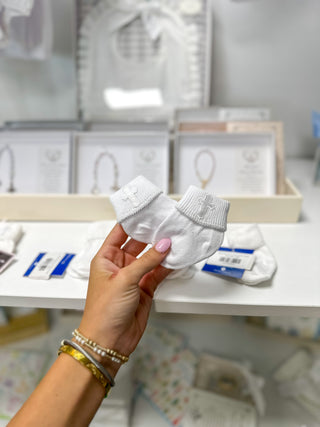 A hand with pink nail polish and gold bracelets holds a pair of white knit baby booties with embroidered details, set against a blurred boutique background.