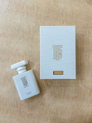 A white rectangular perfume bottle with a matching white box, both featuring a gold monogram logo and a small gold plaque